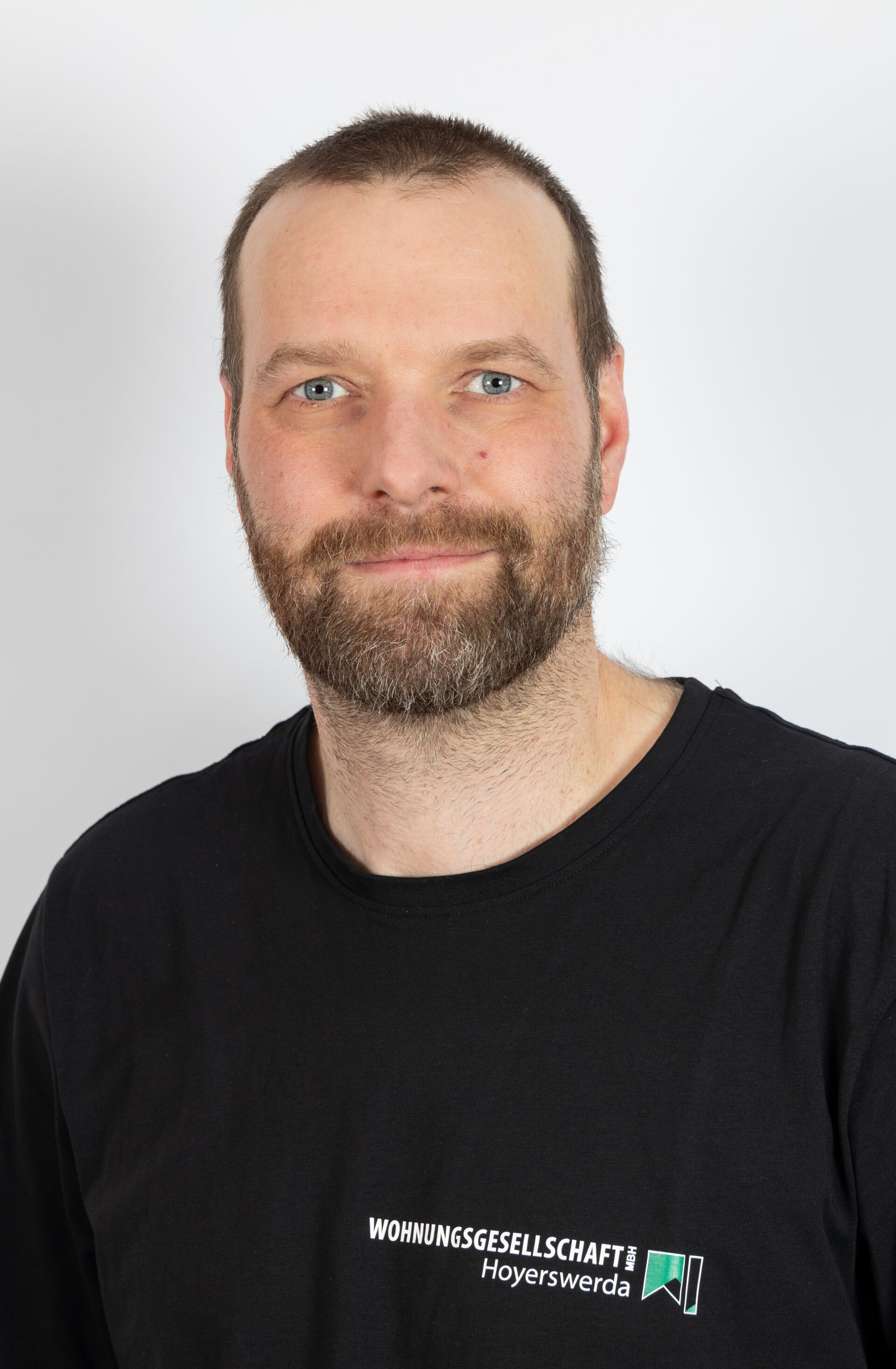 Portraitfoto - Mann mit braunen Haaren, Bart & schwarzem Shirt mit Logo der Wohnungsgesellschaft Hoyerswerda