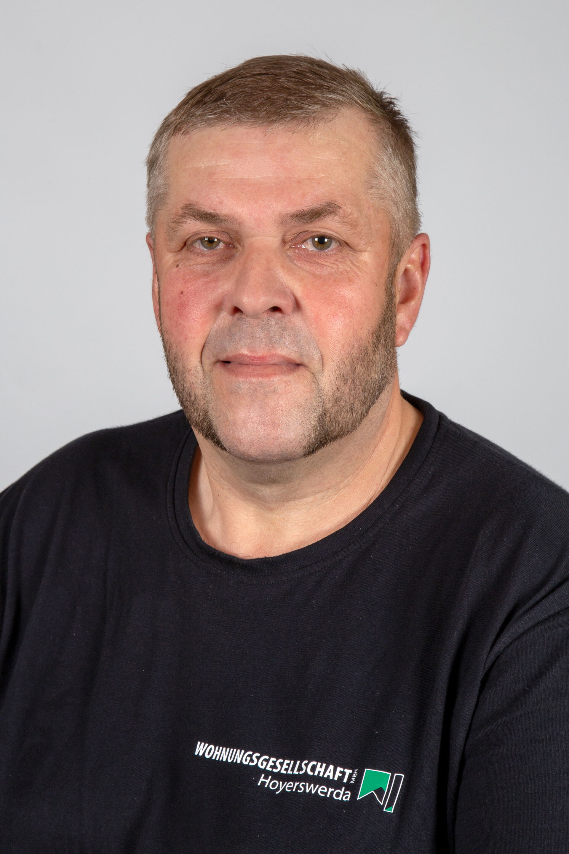 Portraitfoto - Mann mittleren Alters mit graumeliertem Haar & Drei-Tage-Bart in T-Shirt mit Firmenlogo