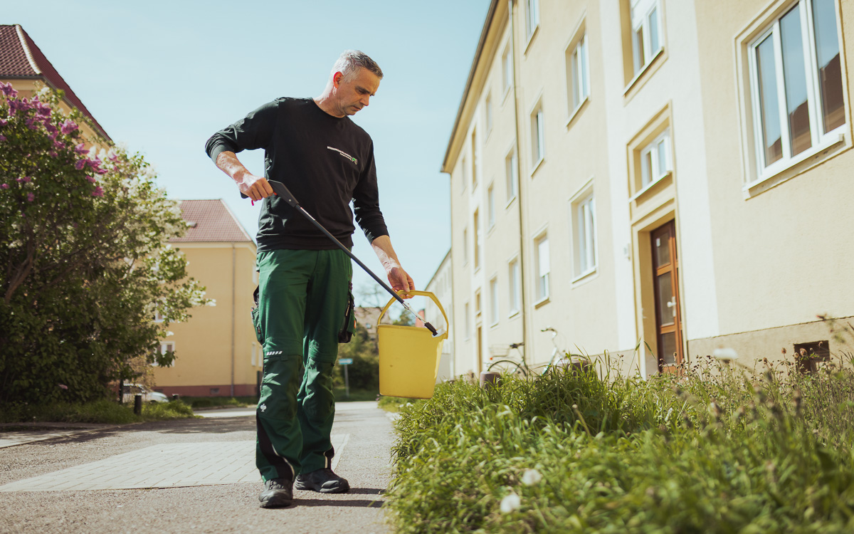 Ein Hausmeister mit gelbem Eimer & Greifzange bei der Grünflächenpflege