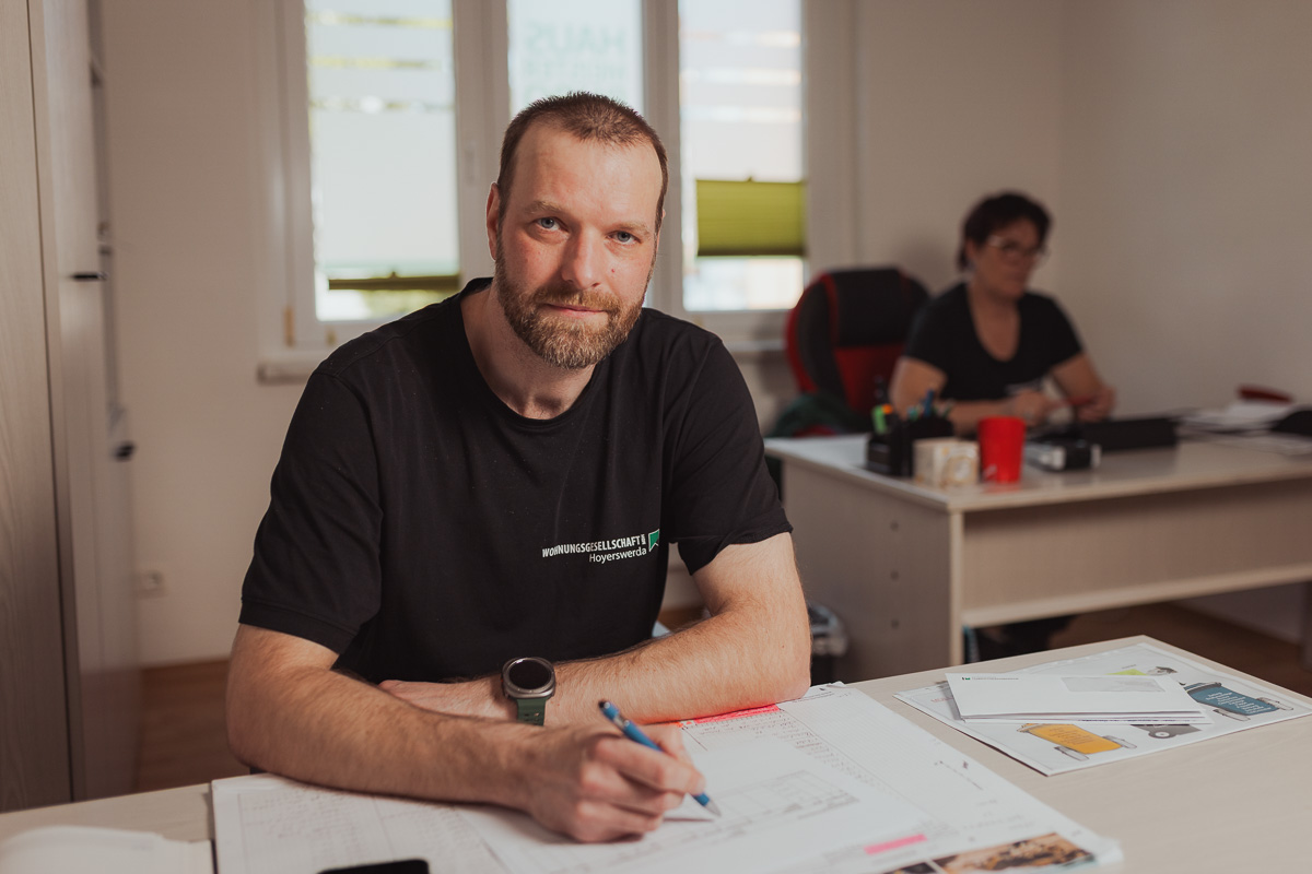 Hausmeister am Schreibtisch bei der Büroarbeit - im Hintergrund sitzt eine Kollegin