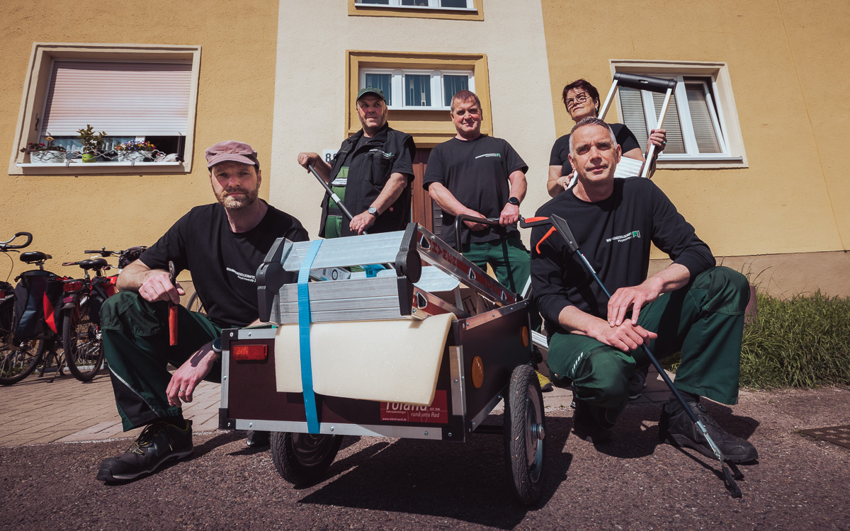 Teamfoto mit fünf Hausmeistern & einem Leiterwagen vor einem gelben Wohnhaus